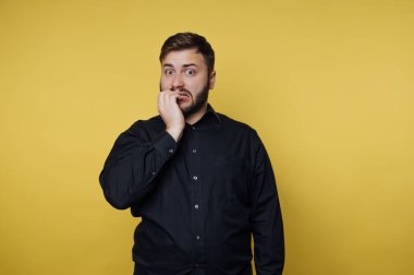 Sarı bir duvarın önünde endişeli bir ifadeyle duran bir adam. Ağzını eliyle kapatıyor. Ortam sıradan bir ortamda kapalı gibi görünüyor..