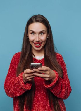 Uzun saçlı bir kadın akıllı bir telefon tutar ve heyecanla gülümser. İçerde gibi görünüyor, saf mavi arka planı ve parlak ışıklandırmasıyla..