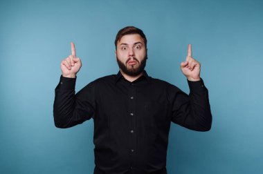 Bir adam mavi bir arka planın önünde duruyor. Siyah bir gömlek giyiyor ve iki eliyle yukarı doğru işaret ediyor. Yüz ifadesi şaşırdığını gösteriyor. Parlak ışıklandırma özelliklerini vurgular.