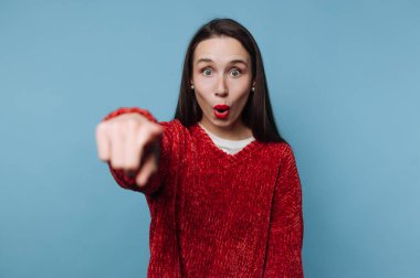 Genç bir kadın arkasında açık mavi bir duvarla içeride duruyor. O, şaşırmış bir bakışla izleyiciyi işaret eder. Kırmızı süveteri arka planda duruyor..