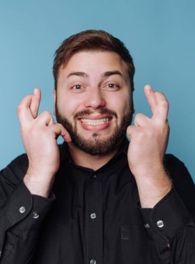 Bir adam mavi bir duvara karşı duruyor, parmakları çapraz ve büyük bir gülümsemeyle. Umutlu görünüyor ve şans getirmesi için jest yaparken doğrudan kameraya bakıyor..