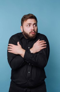 Sakallı bir adam mavi bir arka plana karşı duruyor. Siyah bir gömlek giyiyor ve kollarını kavuştururken şaşırmış görünüyor. Yüzündeki ifade endişe ya da şok gösteriyor..