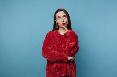 Genç bir kadın sanki düşünüyormuş gibi parmağını dudaklarına koyan neşeli bir ifadeyle ayakta duruyor. O, samimi kırmızı bir kazak giyer. Arkaplan koyu mavi renkte..