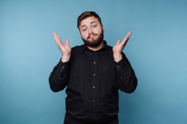 Bir adam elleri havada, kafası karışmış bir ifade sergiliyor. Siyah bir gömlek giyiyor ve mavi arka planlı bir stüdyoda. Bu sahne belirsiz bir anı yakalıyor..