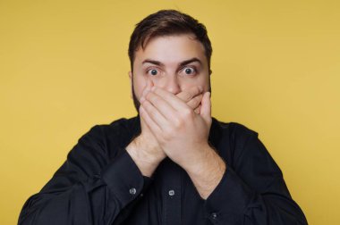 Sarı bir arka planın önünde bir adam duruyor. Onun elleri ağzını kapatıyor. Gözleri açık, gördüklerine şaşırmış ya da şok olmuş..