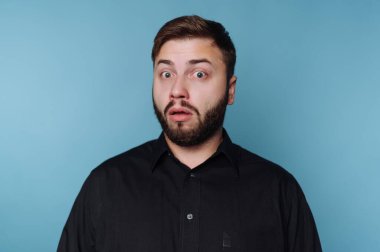 Bir adam basit bir mavi arka planın önünde duruyor. Gözleri ve ağzı açık bir şekilde dikkat çekici bir şaşkınlık ifadesi var. Koyu renk gömleği arka planla çelişiyor..