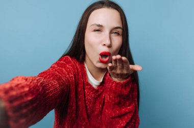 Bir kadının kameraya doğru öpücük üflediği görülüyor. Kırmızı bir kazak giyiyor ve uzun koyu renk saçları var. Arka plan mavi ve ışıklandırma parlak..