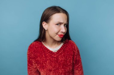 Mavi bir duvarın önünde duran genç bir kadın komik bir ifade takınıyor. Kırmızı bir kazak giyiyor ve yüzünde neşeli bir ifade var..