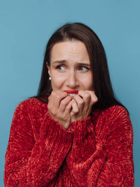 Bir kadın, tırnaklarını yerken ve ellerini ağzına tutarken endişeli bir bakış sergiler. Arka plan basit bir mavi renktir, ifadesini netleştirir ve odaklar..