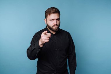 Bir adam mavi bir arka planın önünde duruyor. O ciddi bir ifadeyle izleyiciyi işaret ediyor. Uzun kollu siyah bir gömlek giyiyor ve sakalı var..