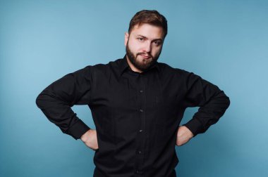 Sakallı bir adam mavi bir duvara karşı poz verir. O, elleri kalçasının üzerinde güvenle duruyor. Yumuşak ışıklandırmalı bir iç mekan gibi görünüyor..