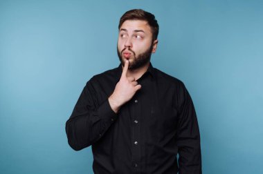 Siyah gömlekli, mavi arka planda duran genç bir adam. Düşünceli bir ifadesi var ve sanki önemli bir şey düşünüyormuş gibi parmağını dudaklarına değdiriyor..