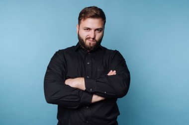 Kolları mavi bir arka planda çapraz duran bir adam. Sakalı var ve kendine güvendiğini ya da meydan okuduğunu gösteren bir ifade takınıyor..