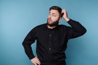 Kafası karışık bir ifadeyle mavi bir arkaplan karşısında duran bir adam. Bir eliyle başını kaşıyor, diğer eli de kalçasında. Kıyafeti sıradan ve basit..