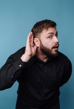 A man leans slightly forward, holding his hand to his ear as if trying to hear someone speak. He wears a black shirt and stands against a blue background.