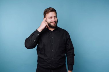 Mavi arka planda duran siyah gömlekli bir adam. O, gülümserken eli başının üzerinde bir jest yapar. Dekor kapalı gibi görünüyor..