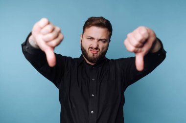 Bir adam mavi bir arka planın önünde duruyor. Yüz kılları var ve siyah bir gömlek giyiyor. Her iki başparmağı da reddediyor. Bu da bir şeyi onaylamadığını gösteriyor..