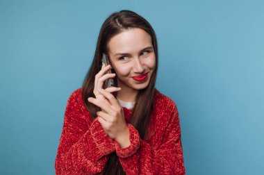 Uzun saçlı bir kadın telefonda konuşurken gülümsüyor. O kırmızı bir kazak giyer. Arka plan mavi ve ayarı parlak görünüyor..