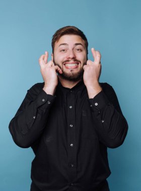 Büyük bir gülümseme ile mavi bir duvara karşı duran bir adam, bir umut jesti olarak parmaklarını çapraz yapar. Siyah bir gömlek giyiyor ve stüdyoda neşeli ve iyimser görünüyor..