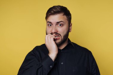 Genç bir adam sarı bir arka planın önünde duruyor. Tırnaklarını yiyor ve endişeli görünüyor. Işıklandırma parlak ve yüz ifadesini vurguluyor. Siyah bir gömlek giyiyor..