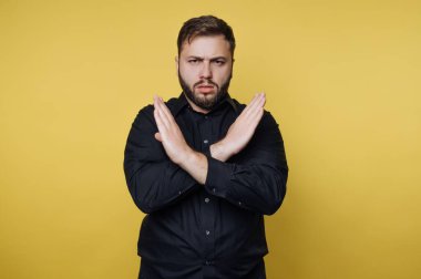 Kollarını vücudunun önünde kavuşturmuş bir adam. Yüz ifadesi ciddiyetini gösteriyor. Sarı duvarlı, parlak kapalı bir alanda siyah bir gömlek giyiyor..