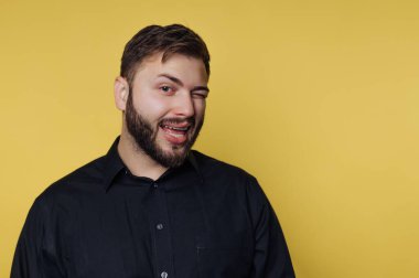 Sarı arka planda bir adam duruyor. Tek gözüyle göz kırpıyor ve gülümsüyor. Siyah bir gömlek giyiyor ve sakalı var. İfadesi eğlenceli ve çekici..