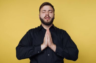 Bir adam gözleri kapalı ve elleri bir dua jestinde birbirine bastırılmış olarak duruyor. Koyu renk bir gömlek giyiyor ve sakalı var. Parlak sarı arka plan onu çevrelemiş..