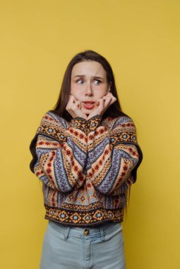 Bir kadın parlak sarı bir duvara karşı duruyor. O, elleri yüzünde endişeli görünüyor. Renkli bir süveter ve kot pantolon giyiyor. Uzun saçları dümdüz dökülüyor..