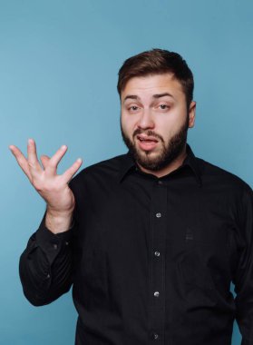 Bir adam içeride mavi bir arka plana karşı duruyor, bir jestle elini kaldırıyor. Sakalı var ve siyah bir gömlek giyiyor. Yüz ifadesi bir şey açıkladığını gösteriyor..