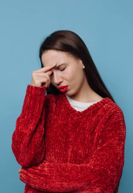 Genç bir kadın bir eliyle alnını tutarken diğer kolu çaprazdır. Kırmızı bir kazak giyiyor ve yorgun görünüyor. Mavi arka plan şu anda onun ruh haline ekleniyor..