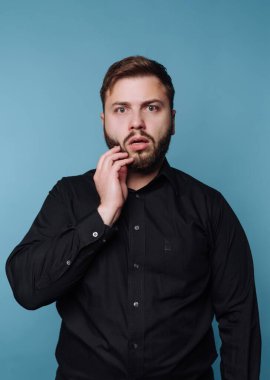 Bir adam mavi bir duvarın karşısında şaşırmış bir şekilde duruyor. Kısa saçlı ve sakallı, siyah bir gömlek giyiyor. Eli yüzüne dokunur, şaşırır ya da şok geçirir..