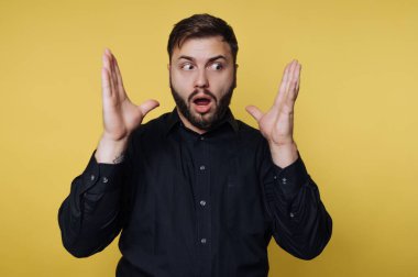 Kapalı bir yerde yüzünde şaşkın bir ifadeyle bir adam duruyor. Beklenmedik bir şeye tepki veriyormuş gibi ellerini kaldırıyor. Arka plan sarı..