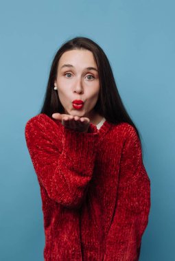 Uzun saçlı bir kadın öpücükle neşeli bir jest yapar. Gülümsüyor ve kırmızı bir kazak giyiyor. Arkaplan koyu mavi renkte, parlak bir sahne oluşturuyor..