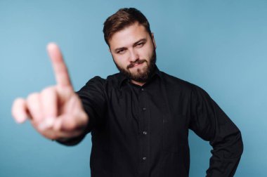 Sol elini kaldırmış, bir parmağını öne doğru gösteren bir adam. Sakalı var ve siyah bir gömlek giyiyor. Arkaplan koyu mavi renkte..