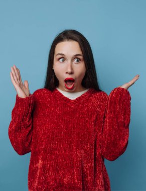 Kırmızı kazak giyen bir kadın sürprizle tepki verir. Mavi bir arka plana karşı duruyor, elleri havada ve yüzünde sevinç ifadesi var..