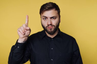 Bir adam parlak sarı bir arkaplanın önünde duruyor. O, siyah bir gömlek giyer ve neşeli bir yüz yaparken bir parmağını kaldırır. Yüzündeki ifade önemli ya da komik bir şey paylaşmak üzere olduğunu gösteriyor..