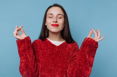 Kırmızı kazak giyen bir kadın gözleri kapalı ve gülümseyerek farkındalık alıştırması yapıyor. Ellerini meditasyon pozisyonunda tutarken rahatlamış görünüyor. Arka plan mavi..