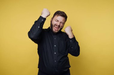 Elleri havada ve sarı bir duvara karşı neşeli bir ifadeyle duran bir adam. Başarılı ya da neşeli bir anı kutluyor gibi görünüyor..