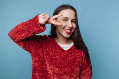 Mavi bir duvarın önünde genç bir kadın duruyor. Gülümsüyor ve barış işareti yapmak için elini kaldırıyor. Kırmızı bir kazak giyiyor ve mutlu görünüyor..