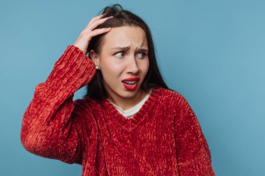Uzun saçlı bir kadın endişeli görünürken elini başına kaldırır. O kırmızı bir kazak giyer ve bir kafa karışıklığı ifadesi gösterir. Arka plan mavi..
