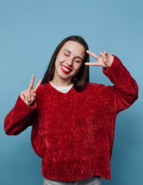 Genç bir kadın, elleri barış işaretiyle mavi bir zemin üzerinde duruyor. Onun uzun siyah saçları ve kırmızı bir kazak giyerken geniş bir gülümsemesi var..