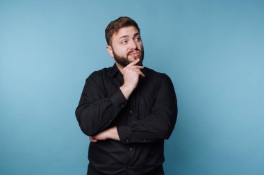 Mavi bir duvarın önünde duran siyah gömlekli bir adam. Düşünceli bir yüz ifadesi var. Düşüncelere dalmış, başını yan tarafa çevirirken çenesini dinlendiriyor..