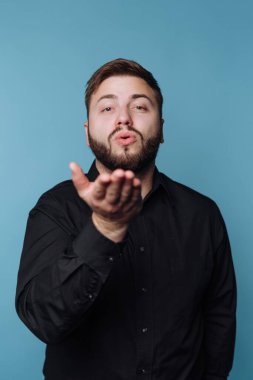 Kameraya öpücük gönderen bir adam görülüyor. Düşünceli bir ifadesi var ve siyah bir gömlek giyiyor. Arkaplan, çarpıcı bir karşıtlık yaratan koyu mavi bir renktir..