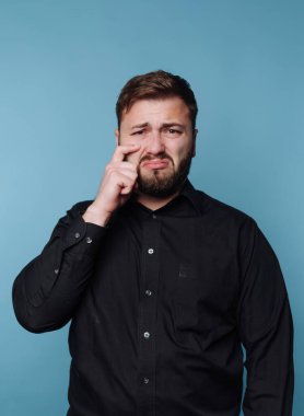 Bir adam mavi bir duvara yaslanır ve üzgün bir yüz gösterir. O, aşağı bakarken bir eliyle gözüne dokunur. Gömleği siyah ve kapalı bir alanda gibi görünüyor..