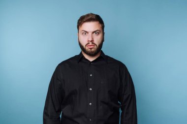 Mavi bir duvarın önünde duran bir adam şaşırmış bir bakış sergiliyor. Siyah bir gömlek giyiyor ve sakalı var. Onun ciddi ifadesi spontane bir anda dikkat çeker..