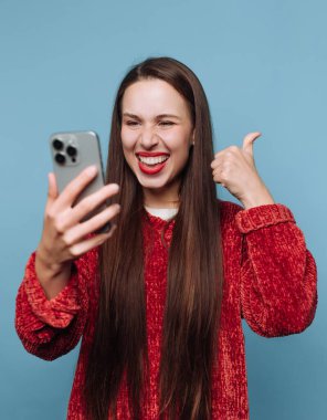 Uzun saçlı bir kadın akıllı telefonuna gülümserken başparmağını kaldırıyor. Kırmızı bir kazak giyiyor ve yüzünde mavi bir duvara karşı neşeli bir ifade var..