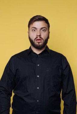 Büyük gözleri ve ağzı açık duran bir adam, kapalı bir alanda şaşkınlık bakışları sergiliyor. Siyah bir gömlek giyiyor ve arkasında düz sarı bir duvar olan bir sakalı var..