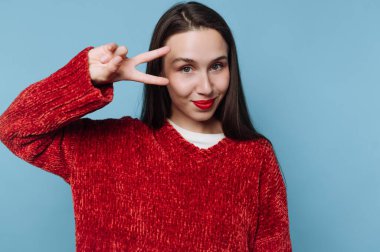 Genç bir kadın mavi bir arka planın önünde duruyor. Kırmızı bir kazak giyiyor ve gülümserken sağ eliyle barış işareti yapıyor. Ayar aydınlık ve neşeli..