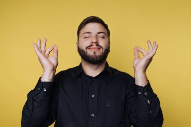 Sakallı bir adam gözleri kapalı oturur, meditasyonu göstermek için el hareketleri yapar. Arka plan parlak sarı, siyah elbisesiyle koyu bir kontrast oluşturuyor..