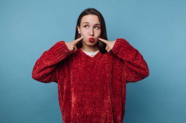 Genç bir kadın mavi bir duvara karşı içeride duruyor. Kırmızı bir kazak giyiyor ve iki parmağıyla yanaklarını göstererek komik bir yüz ifadesi takınıyor. Bu an onun neşeli ruhunu yakalıyor..